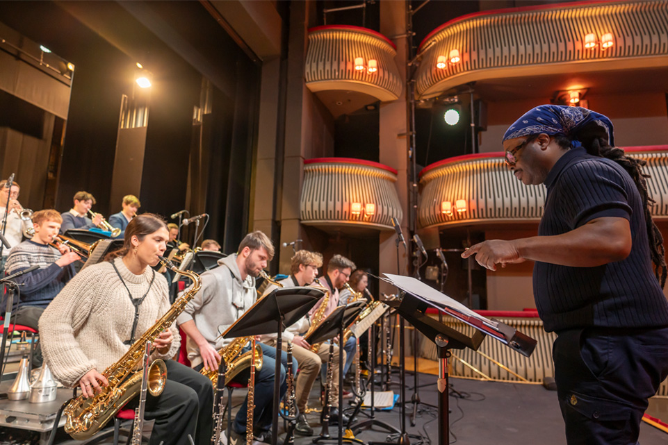 A male conductor, wearing a hair wrap, wearing a dark blue shirt, conducting a jazz orchestra on a stage, with a group of saxophones performing in the front row.